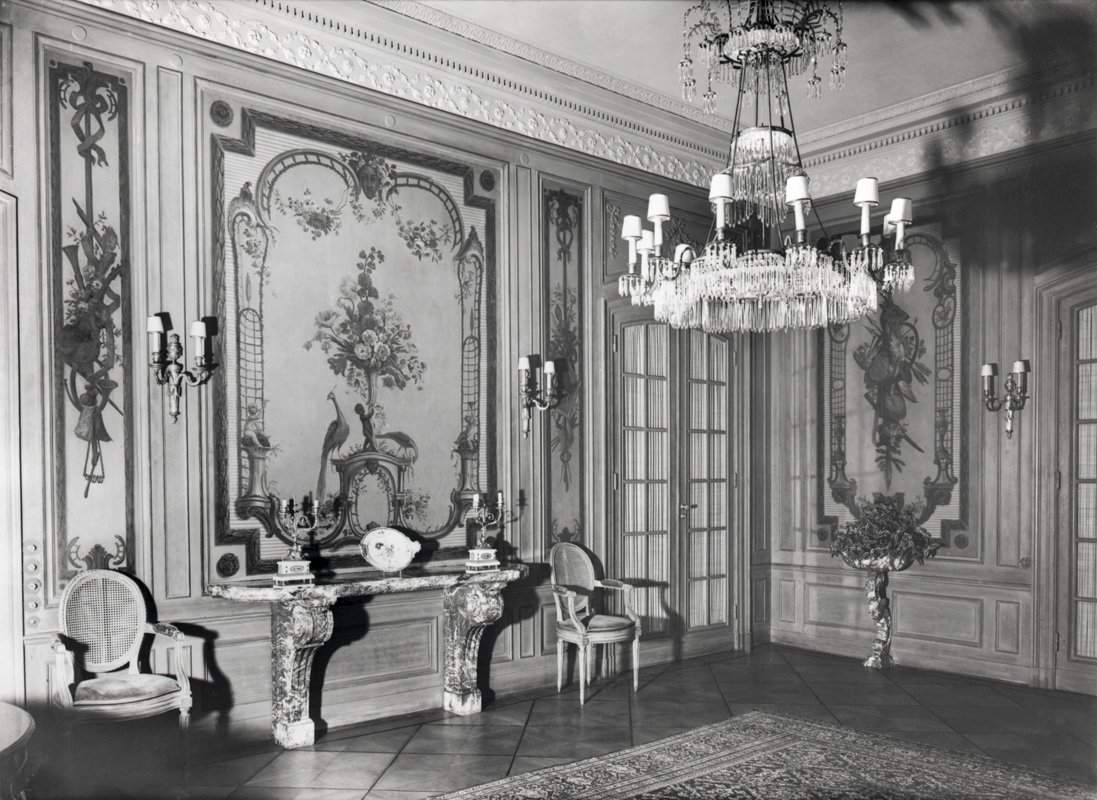 Ein Raum mit viel Schmuck, von der Decke hängt ein Kronleuchter, es gibt Wandmalereien. An der Wand stehen zwei Stühle. Das Foto ist schwarz-weiß.