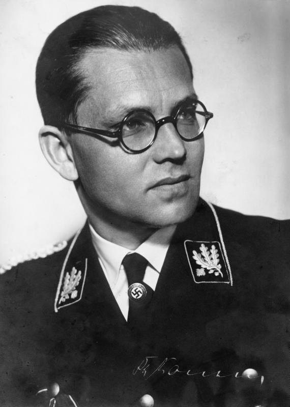 Philipp Bouhler trägt auf dem Foto eine Nazi-Uniform. Seine Haare sind an den Kopf gekämmt, er hat eine Brille auf. Das Foto ist schwarz-weiß.