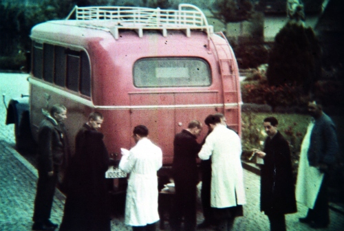 Im Hintergrund ein Bus, davor Patienten, die in den Bus gebracht werden. Ärzte in weißen Kittel tragen sie auf einer Liste ein.