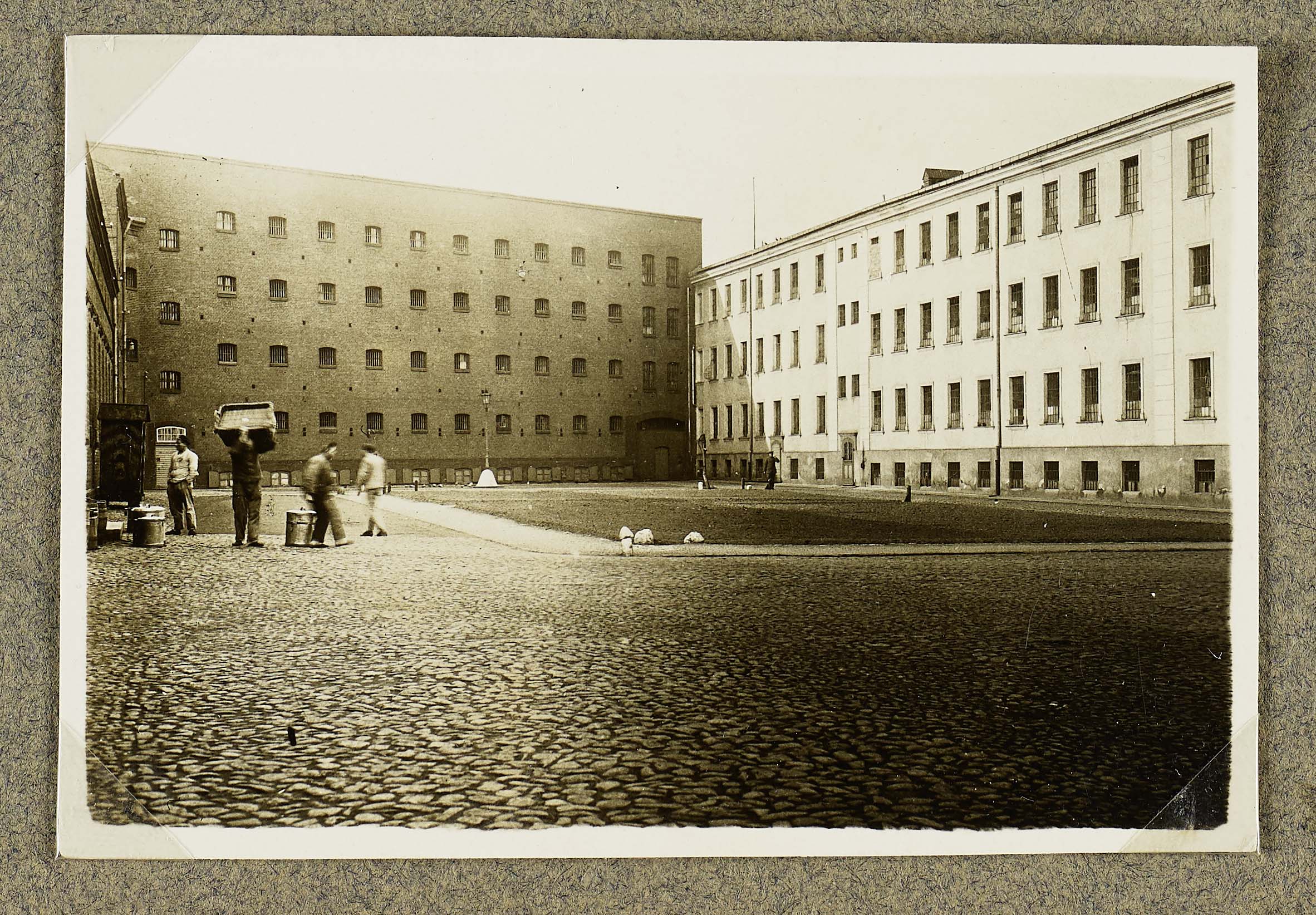 Der Gefängnishof. Er wird umrahmt von hohen Zellengebäuden und einem Wirtschaftsgebäude auf der linken Seite. Links im Vordergrund entladen Häftlinge etwas. Das Foto ist schwarz-weiß.