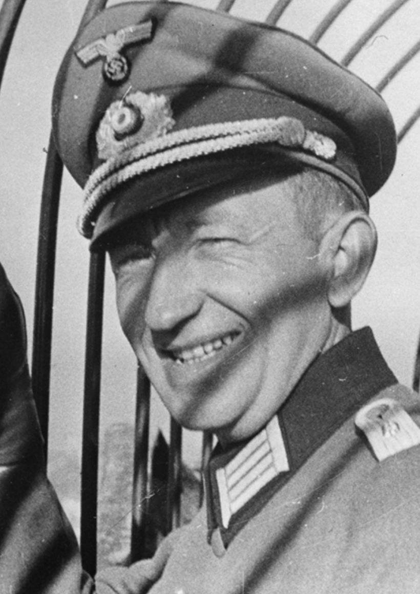 Aquilin Ulrich trägt eine Nazi-Uniform mit Mütze. Seine Hand umfasst eine Metallkonstruktion. Ein Schatten ist auf seinem Gesicht. Das Foto ist schwarz-weiß. 