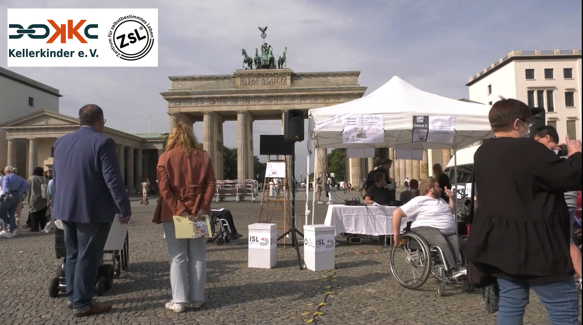 Eine Pavillionzelt, darin ein Tisch mit Technik und ein Infostand. Davor Menschen; im Hintergrund das Brandenburger Tor. Das Foto ist in Farbe.