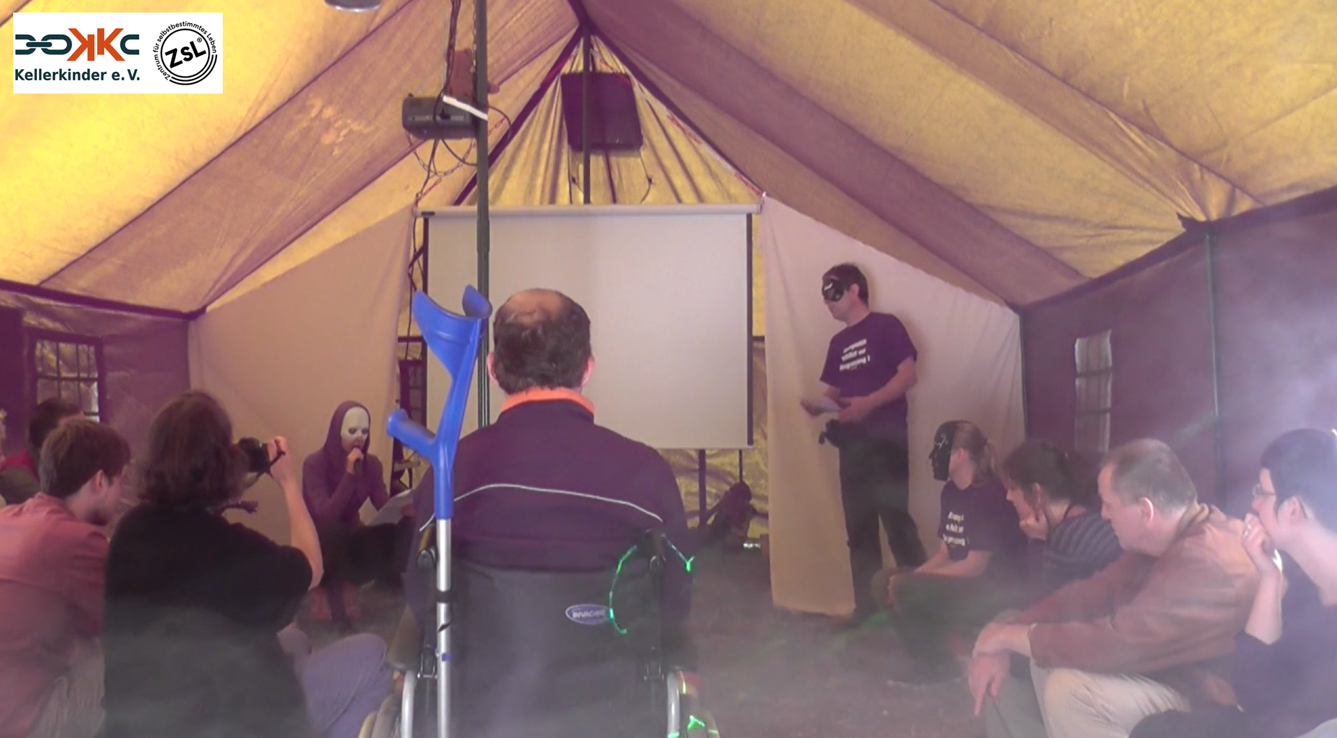 Das Innere eines Zeltes, darin ca. 12 Personen, einige tragen Masken. Eine Person hält ein Mikrofon und spricht die anderen hören zu. Das Foto ist in Farbe.