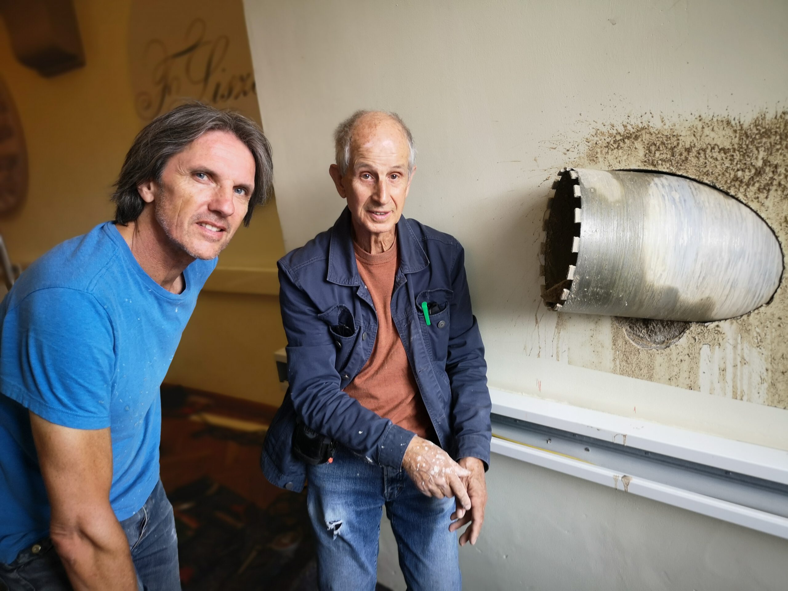 Der Mann links ist Andreas Knitz. Er hat halblange Haare und trägt ein blaues T-Shirt. Der Mann in der Mitte ist Horst Hoheisel. Er hat kurze Haare. Andreas und Horst stehen neben einem Rohr. Das Rohr kommt aus einer Wand. Es ist Teil von einem Kunstwerk. Das Foto ist in Farbe.