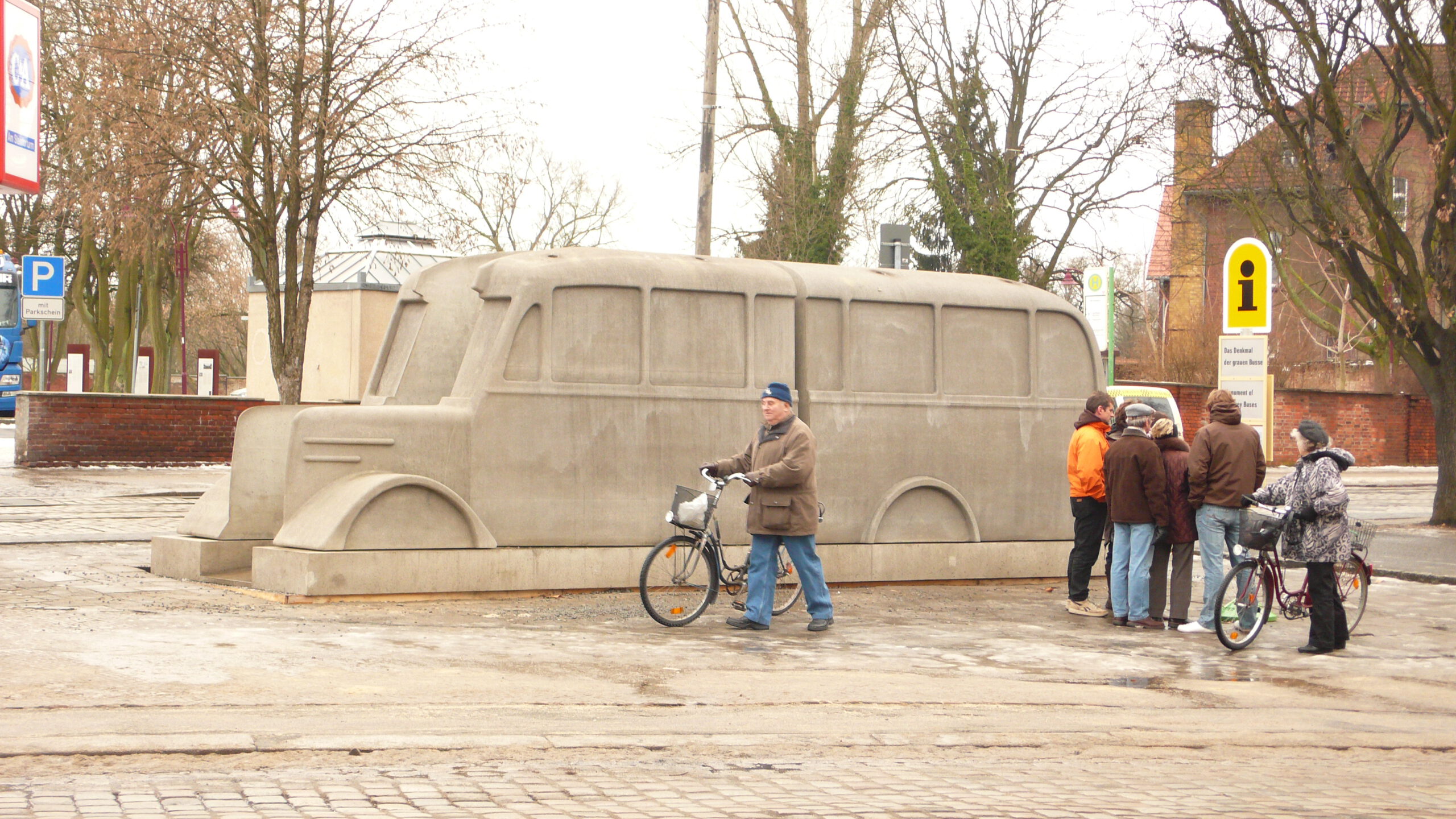 Das Denkmal der Grauen Busse auf dem Nicolaiplatz in Brandenburg an der Havel. 5 Personen lesen den Text auf der Informationstafel. Ein Mann und eine Frau schieben ihr Fahrrad vorbei. Das foto ist in Farbe.