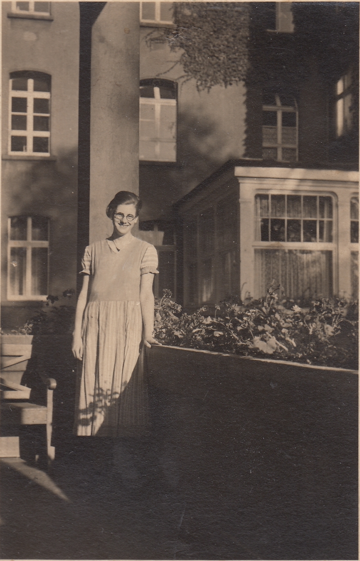 Anna lächelt. Sie trägt ein Kleid und eine Brille. Im Hintergrund ein Gebäude. Rechts neben Anna ein erhöhtes Beet. Auf dessen Umgrenzung stützt sie ihre Hand. Das Foto ist schwarz-weiß.